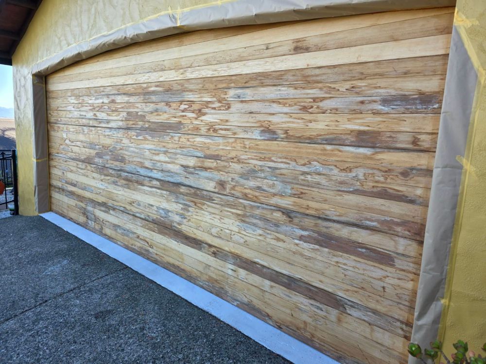 A timber garage door stripped back to bare, blotchy wood with the surrounding wall masked off
